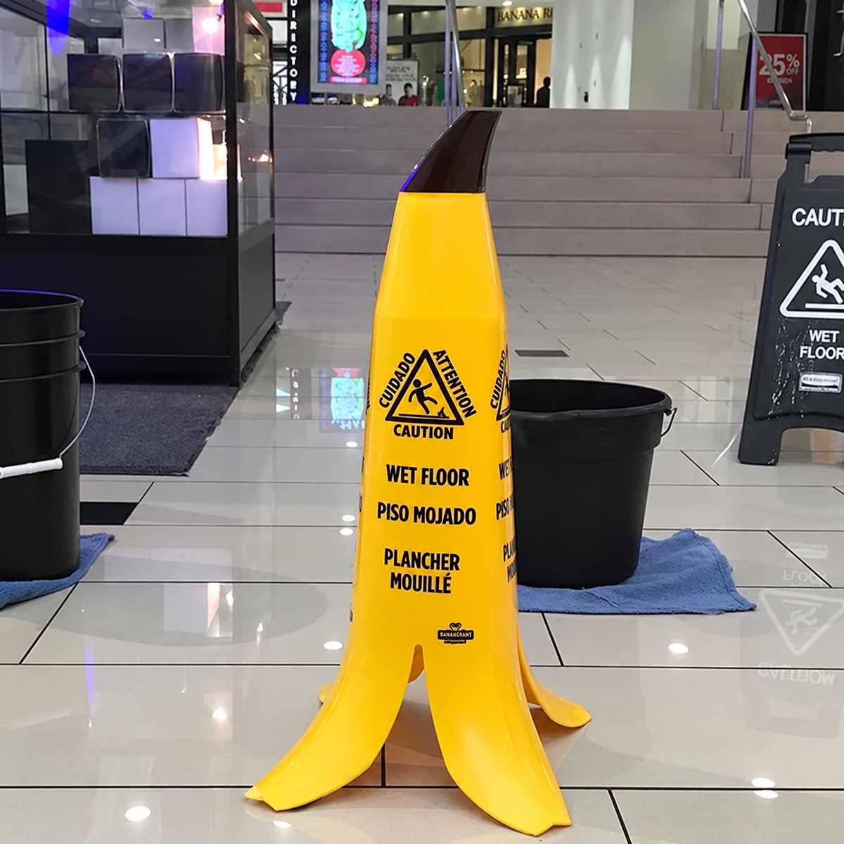 A novelty wet floor sign shaped like a banana peel stands in a tiled indoor mall or lobby area. The sign is bright yellow with black printed warning text and symbols, and black buckets sit on blue cloths near it.